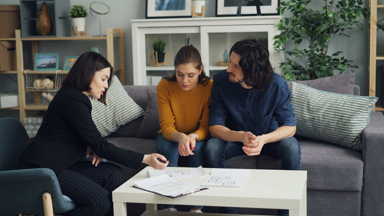 A real estate agent discusses house plans with a couple in a modern living room setting.