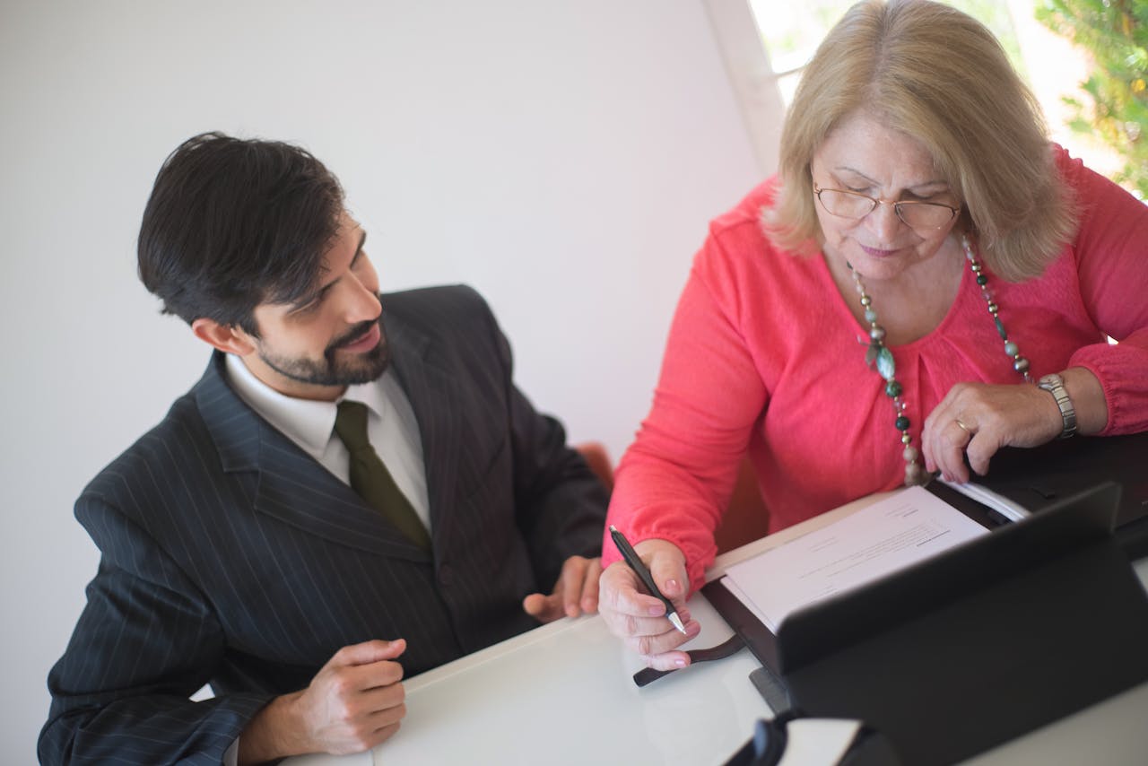 A senior woman signing paperwork with a young professional assisting her at a desk.